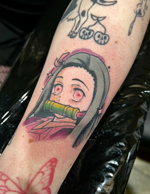 Colorful anime-style tattoo on arm featuring character with wide eyes and yellow pipe in mouth, wearing gray hood