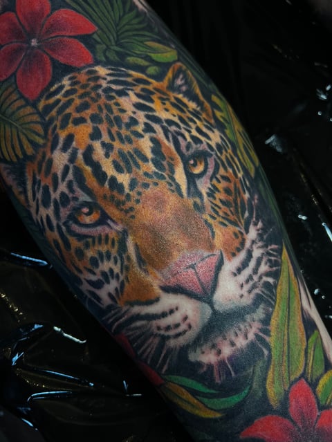 Detailed tattoo of a leopards face with spotted pattern, surrounded by red flowers and green leaves on a persons arm