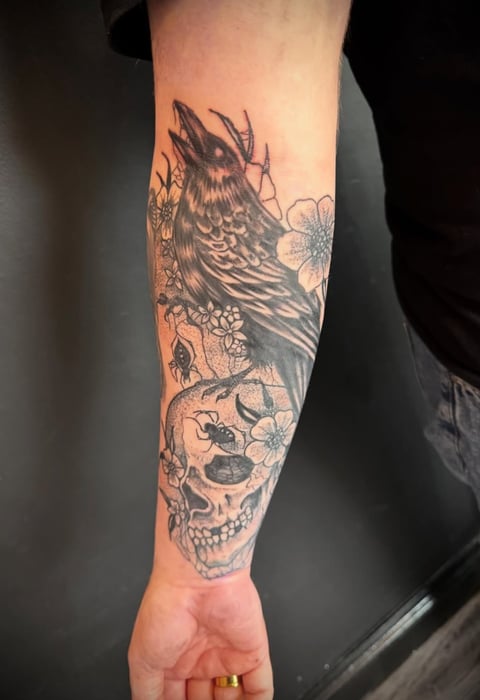 Detailed black ink tattoo on forearm featuring a raven perched above a skull with floral designs and botanical elements