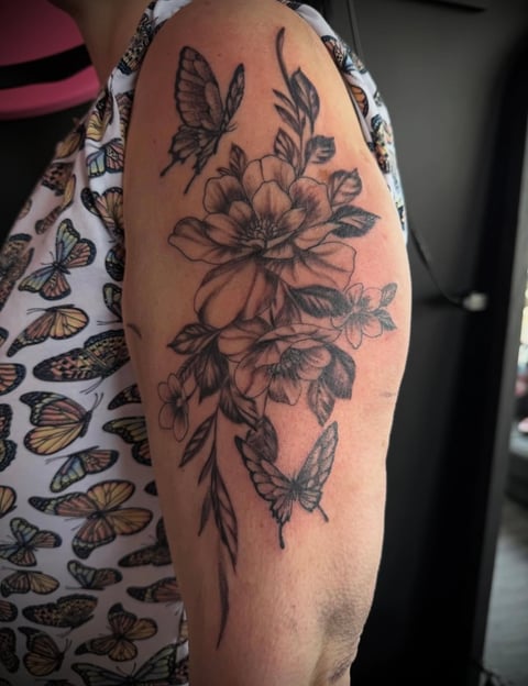 Tattoo on upper arm featuring detailed black ink design of flowers, butterflies, and botanical elements arranged vertically