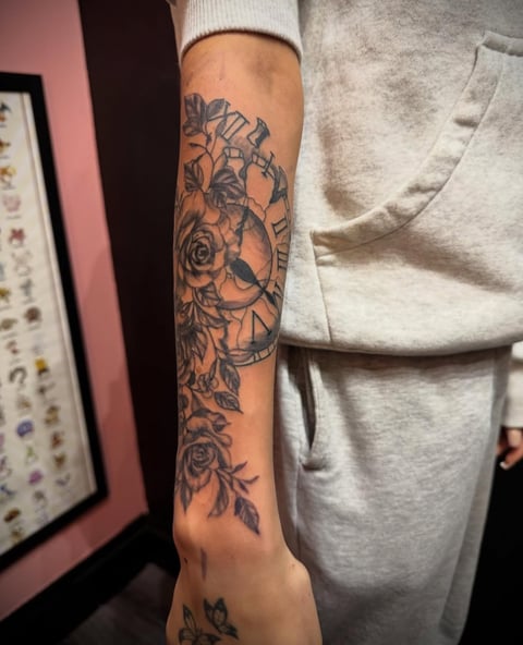 Person wearing light gray hoodie displaying a detailed black floral and clock tattoo on their forearm