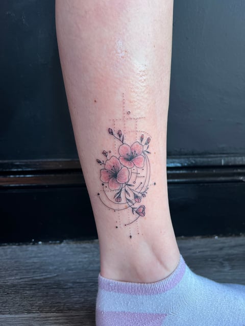 Delicate floral tattoo on lower leg featuring pink flowers with geometric and celestial design elements
