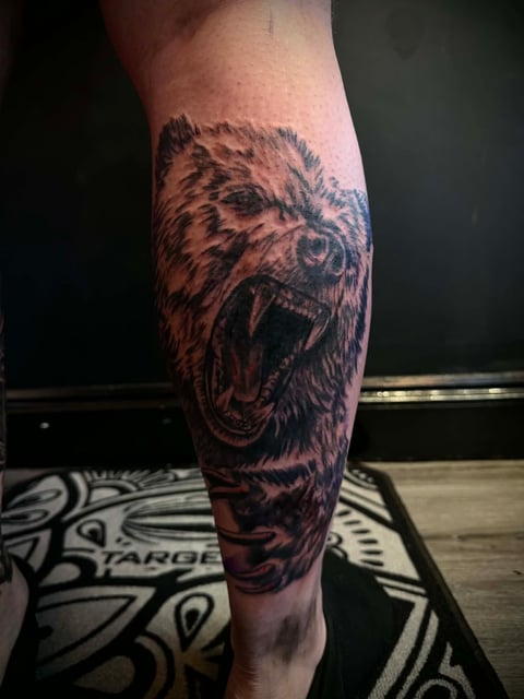 Detailed tattoo of a roaring bear's head on a person's calf, rendered in black and gray ink with intricate shading and detail