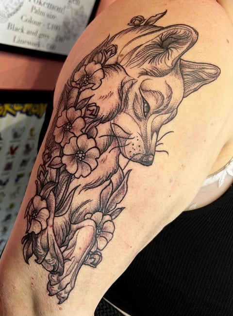 Black ink tattoo on upper arm featuring a fox head with flowers flowing down the side in detailed line art style