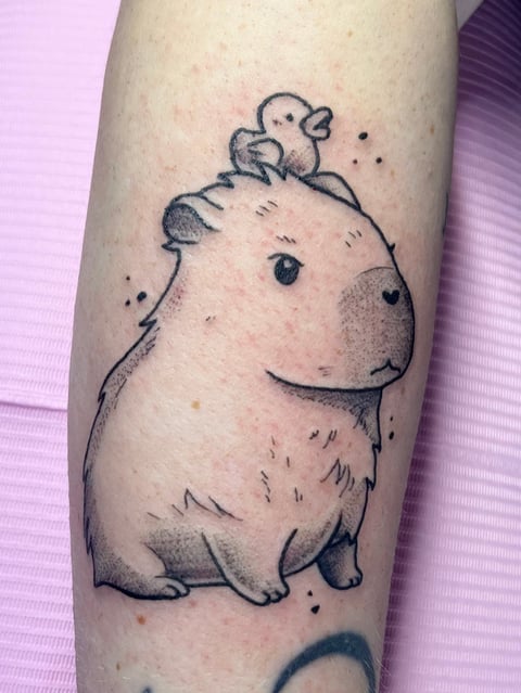 Tattoo of a chubby capybara with a small bird perched on its head on someones arm