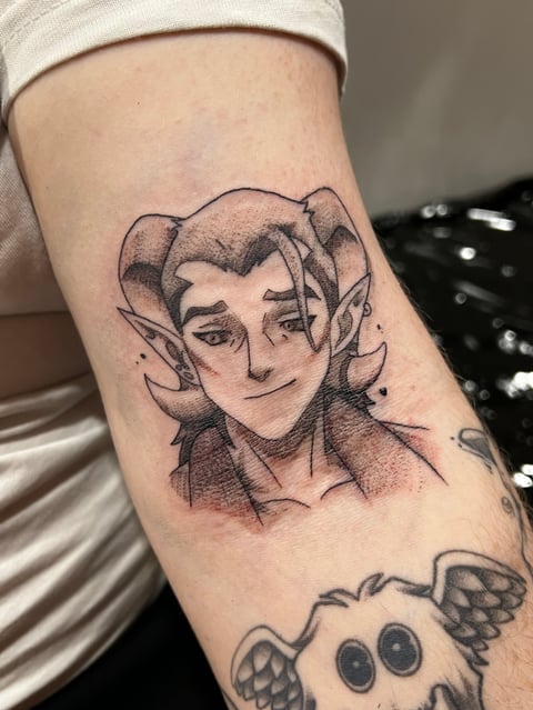 Tattoo of a stylized character with wavy hair and distinctive features on an arm, with another small inked design below